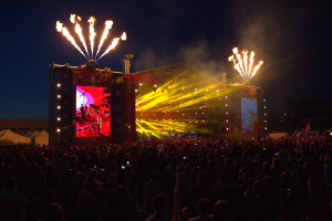 Eine große Menschenmenge auf einem Musikfestival mit einer hell erleuchteten Bühne, auf der Musiker spielen, Banner und Zelte links und Feuerwerk am dunklen Himmel.
