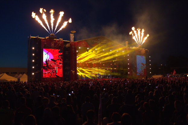 Eine große Menschenmenge auf einem Musikfestival mit einer hell erleuchteten Bühne, auf der Musiker spielen, Banner und Zelte links und Feuerwerk am dunklen Himmel.