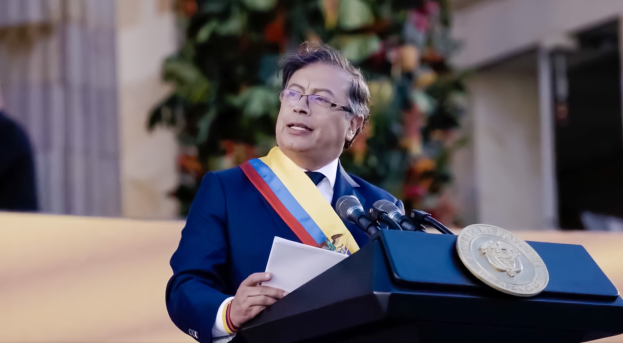 Ein Mann in einem blauen Anzug und Krawatte steht an einem Rednerpult, hält Papiere in der Hand und spricht in ein Mikrofon, mit einer Pflanze und einer Wand im Hintergrund, was auf eine formelle Umgebung hindeutet.