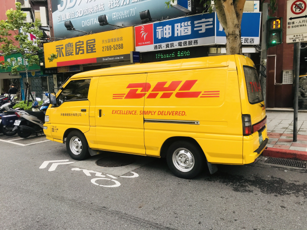 Gelbes DHL-Lieferfahrzeug auf einer Straße neben Gebäuden, Bäumen, Texttafeln, Pfählen, Verkehrsampeln und einer Person auf dem Gehweg geparkt.