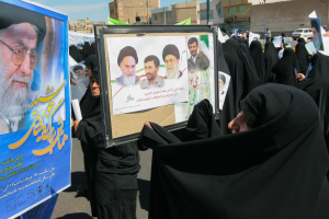 Eine Gruppe von Frauen in schwarzen Abayas hält Plakate mit Fotos von Ajatollah Ali Khamenei hoch, mit Gebäuden, Pfählen, Fahrzeugen und einem klaren blauen Himmel im Hintergrund.