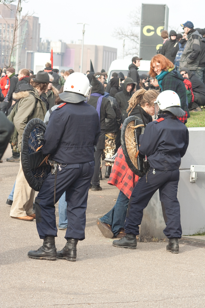 Eine Gruppe von Menschen, die auf einer Straße gehen, mit zwei Personen in der Mitte, die wie Polizisten aussehen, Gebäuden im Hintergrund und Boden unten.