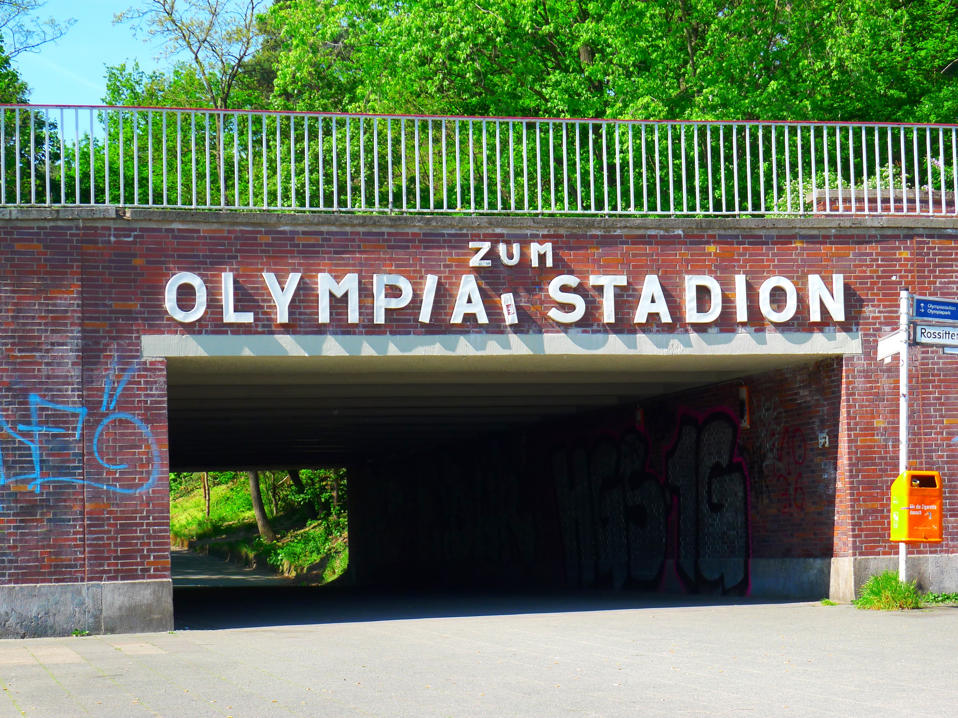 Der Eingang zum Olympiastadion in Berlin, Deutschland, mit einer Brücke mit Text, einem Metallzaun, einem Schild, einer Box, Pflanzen, Gras, einer Baumgruppe und einem bewölkten Himmel.