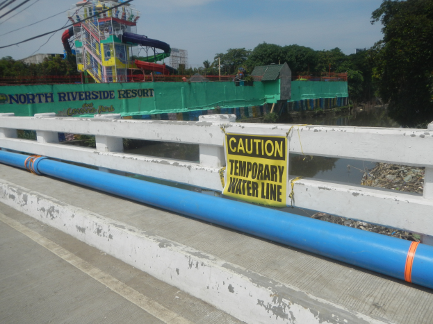 Eine Brücke mit einem "Vorsicht temporäre Wasserleitung"-Schild, Geländer, Bäumen, Gebäuden, einer Wasserrutsche und einem bewölkten Himmel im Hintergrund.