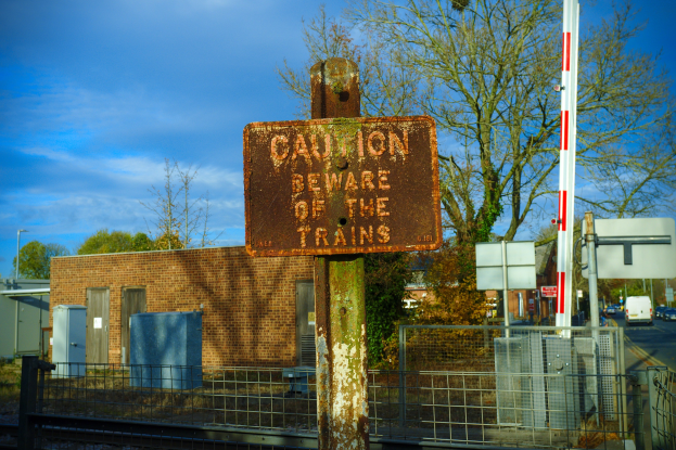 Warnschild an einer Bahnschiene neben einem Metallzaun, mit Bäumen, Masten, einem Gebäude mit Fenstern, einigen Containern, einer Straßenlaterne, einigen Fahrzeugen auf der Straße und einem bewölktem Himmel.