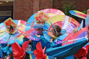 Eine Gruppe von Kindern in bunten Kostümen fährt auf einem Paradewagen, mit Schirmen, mit einem Gebäude, Pflanzen und Lichtern im Hintergrund.