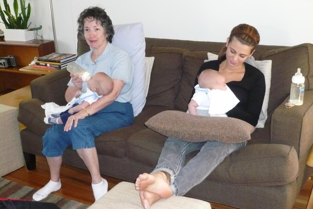 Zwei Frauen sitzen auf einem Sofa und halten Babys, mit Kissen drumherum, einer weißen Wand im Hintergrund und einem Schreibtisch mit Büchern und einer Zimmerpflanze links daneben.