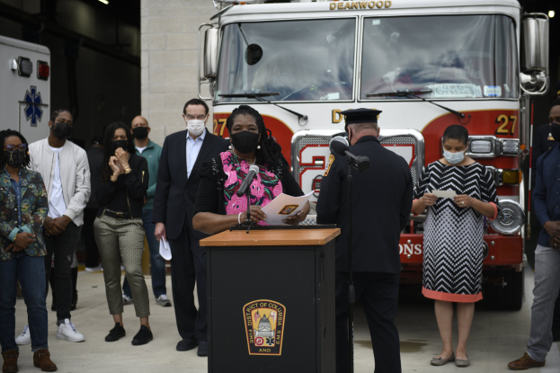 Bürgermeisterin Lori Lightfoot spricht bei einer Pressekonferenz vor einem Feuerwehrauto, umgeben von maskierten Menschen und einer beleuchteten Wand im Hintergrund.