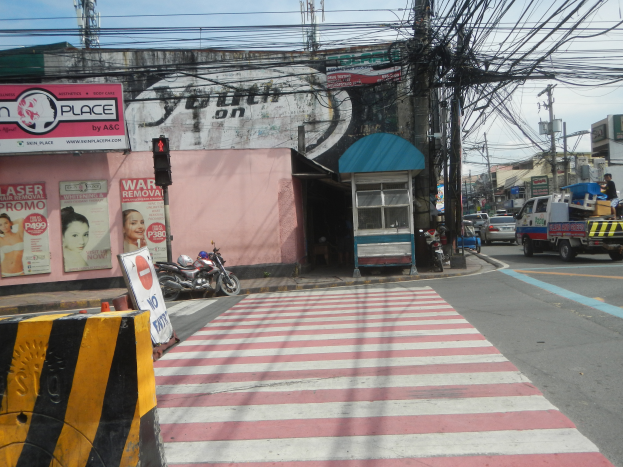 Eine Stadtstraße mit Zebrastreifen, Fahrzeugen, einer Trennlinie, Verkehrsampeln, Schildern mit Text und Bildern, Gebäuden, Strommasten mit Drähten und einem bewölkten Himmel.