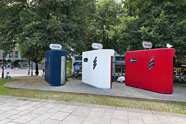Drei mobile Toiletten in einer Reihe auf einer grasbewachsenen Straßenecke mit Gebäuden und einem klaren blauen Himmel im Hintergrund.