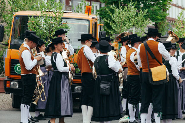 Gruppe von Menschen in traditioneller bayrischer Tracht, die die Straße entlangmarschieren, Instrumente spielen und Taschen tragen, mit Bäumen und Gebäuden im Hintergrund und einem geparkten Fahrzeug.