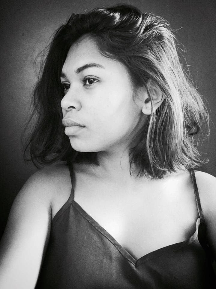 Eine selbstbewusste Frau in einem schwarzen Kleid posiert in einem Schwarz-Weiß-Foto, das vor einer Wand mit einem glatten Bob und natürlichem Make-up steht.