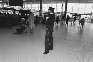 Polizeibeamter in Uniform mit Waffe in einem Flughafen-Terminal umgeben von Menschen, Gepäckwagen und Hintergrund-Flughafen-Strukturen.