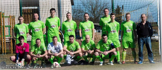 Eine Gruppe von Männern in grünen T-Shirts steht und hockt auf einem Fußballfeld mit einem Ball davor, ein Torpfosten rechts und ein Hintergrund aus Bäumen, Gebäuden, Pfählen, Fahrzeugen und Himmel, mit Text unten links.