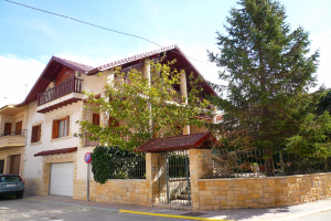 Eine Villa mit einem davor geparkten Auto, das Fenster, Geländer, Pfeiler und ein Dach hat, umgeben von einem Tor, einem Schild, einem Straßenpfahl, Bäumen und einer Wand unter einem bewölktem Himmel.