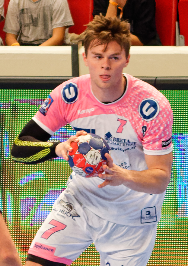 Ein Mann in einem pinken Hemd hält einen Fußball, mit der Hand eines anderen Menschen auf der linken Seite; im Hintergrund sitzen ein paar Menschen auf Stühlen, was auf ein Handballspiel hindeutet.