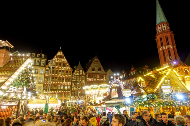 Ein belebter Weihnachtsmarkt in Nürnberg, Deutschland mit Menschen um dekorierte Stände versammelt, Gebäuden mit Fenstern und einem Kirchturm im Hintergrund und einem dunklen Himmel.