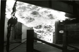 Ein Schwarz-Weiß-Foto eines Feuers in einem Gebäude, mit einer Wand auf der rechten Seite, die eine Uhr zeigt, und einem Boden unten.