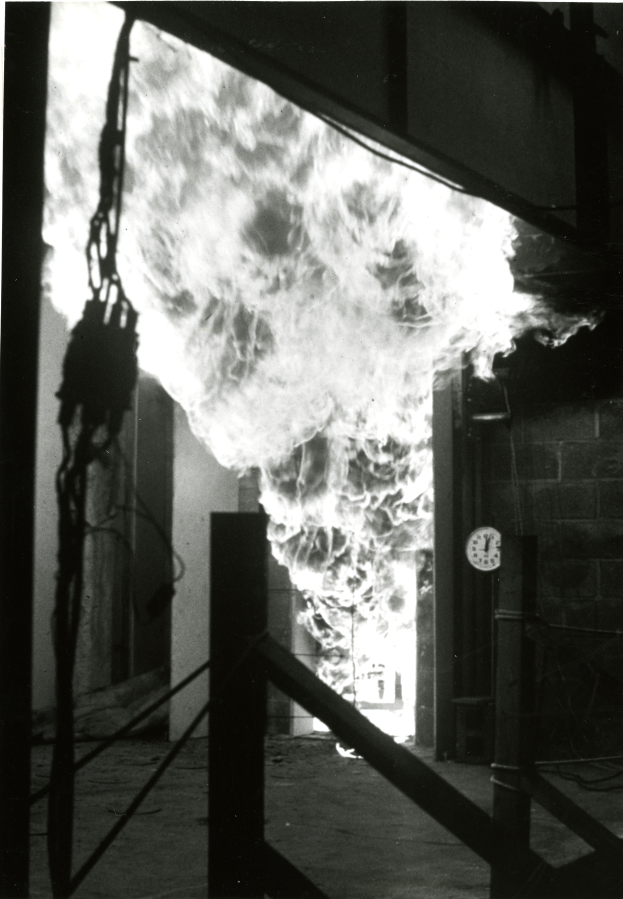 Ein Schwarz-Weiß-Foto eines Feuers in einem Gebäude, mit einer Wand auf der rechten Seite, die eine Uhr zeigt, und einem Boden unten.