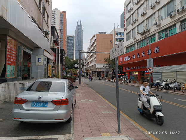 Ein Mann fährt mit einem Roller eine städtische Straße entlang, gesäumt von hohen Gebäuden, mit Fußgängern auf den Bürgersteigen und anderen Fahrzeugen auf der Straße, unter einem klaren blauen Himmel mit Bäumen im Hintergrund.