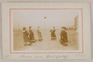 Eine Gruppe von Frauen, die auf dem Boden Füßball spielen, mit einem Ball in der Mitte, Pfählen und Gebäuden im Hintergrund und Text unten auf dem Bild.