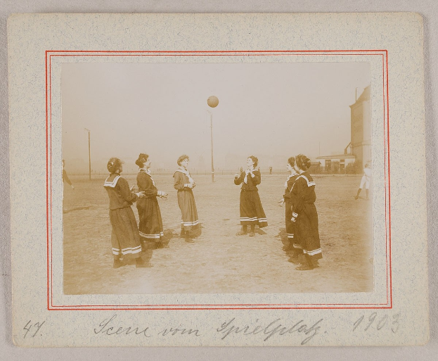 Eine Gruppe von Frauen, die auf dem Boden Füßball spielen, mit einem Ball in der Mitte, Pfählen und Gebäuden im Hintergrund und Text unten auf dem Bild.