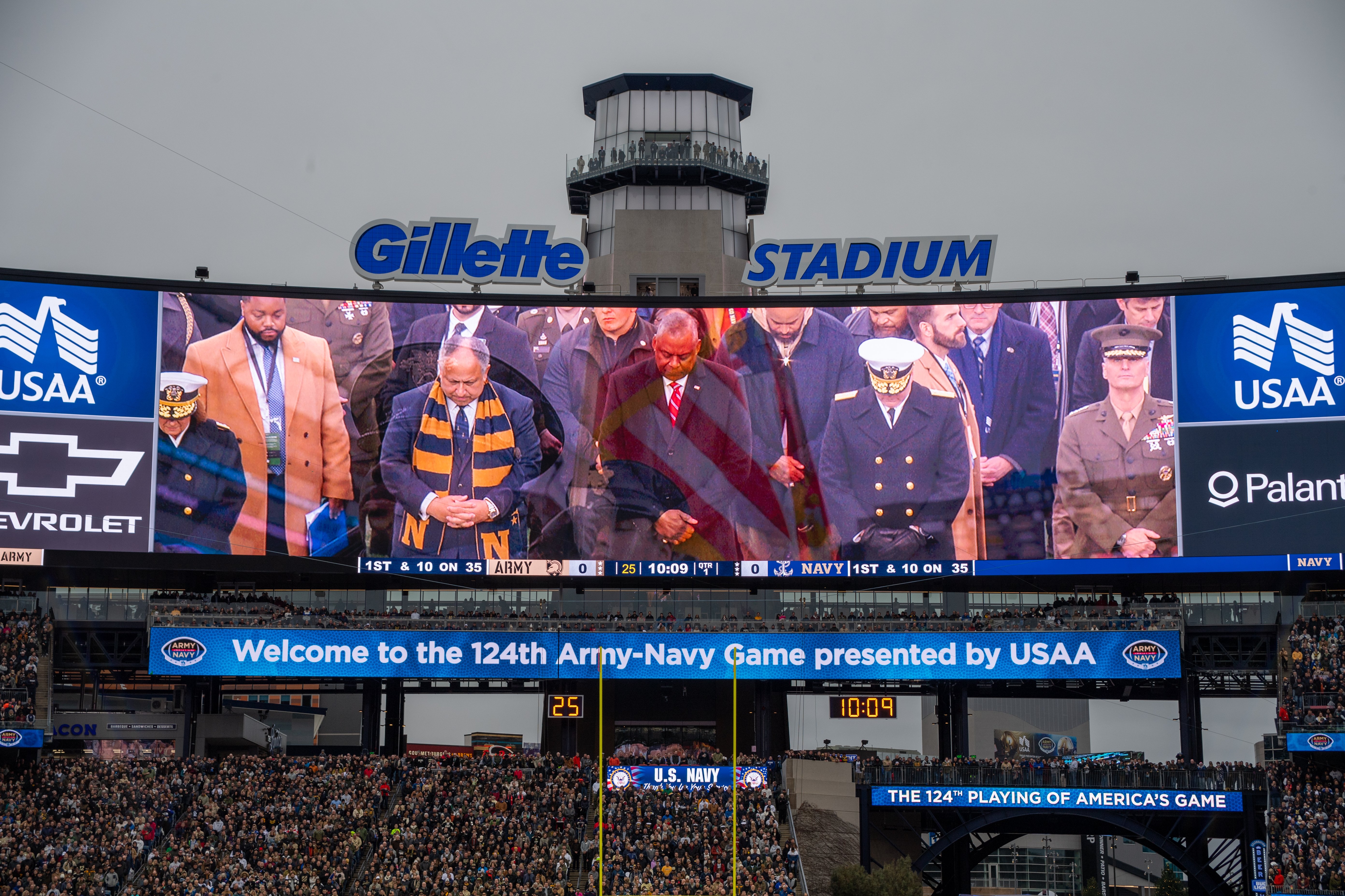 Eine große Menschenmenge in einem Stadion schaut ein Football-Spiel, mit einem zentralen Bildschirm, der Bilder und Text anzeigt, Schilder mit Text, Zuschauern auf den Tribünen, einem Bogen, Bäumen und einem klaren blauen Himmel im Hintergrund.