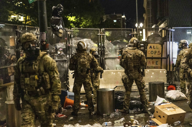 Eine Gruppe von Polizisten in Uniform, Helmen und mit Taschen steht in einer Reihe vor einem Zaun bei Nacht, jeder hält eine Waffe in der Hand. Der Boden ist mit Mülltonnen, Flaschen, Kartons und anderem Müll übersät. Im Hintergrund gibt es Bäume, Gebäude, Laternenpfähle und ein Schild an einem Pfahl.