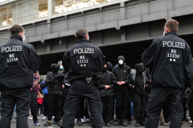 Eine Gruppe von Polizisten in Uniform steht vor einer Menge von Menschen in schwarzen Uniformen und Masken, mit einer Brücke und einem Gebäude im Hintergrund, was eine städtische Umgebung vermuten lässt.