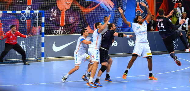 Eine Gruppe von Männern, die Handball auf einem Platz mit einem Netz auf der linken Seite spielen, mit Bannern im Hintergrund und Menschen mit Kameras, die die Aktion aufnehmen.