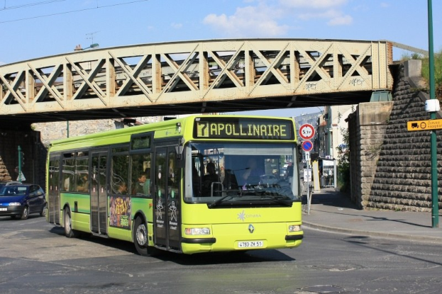 Grüner Bus fährt unter einer Brücke auf einer Stadtstraße mit anderen Fahrzeugen, einem Pfahl mit Schildern, einer Wand, Gebäuden, Bäumen und einem bewölkten Himmel.