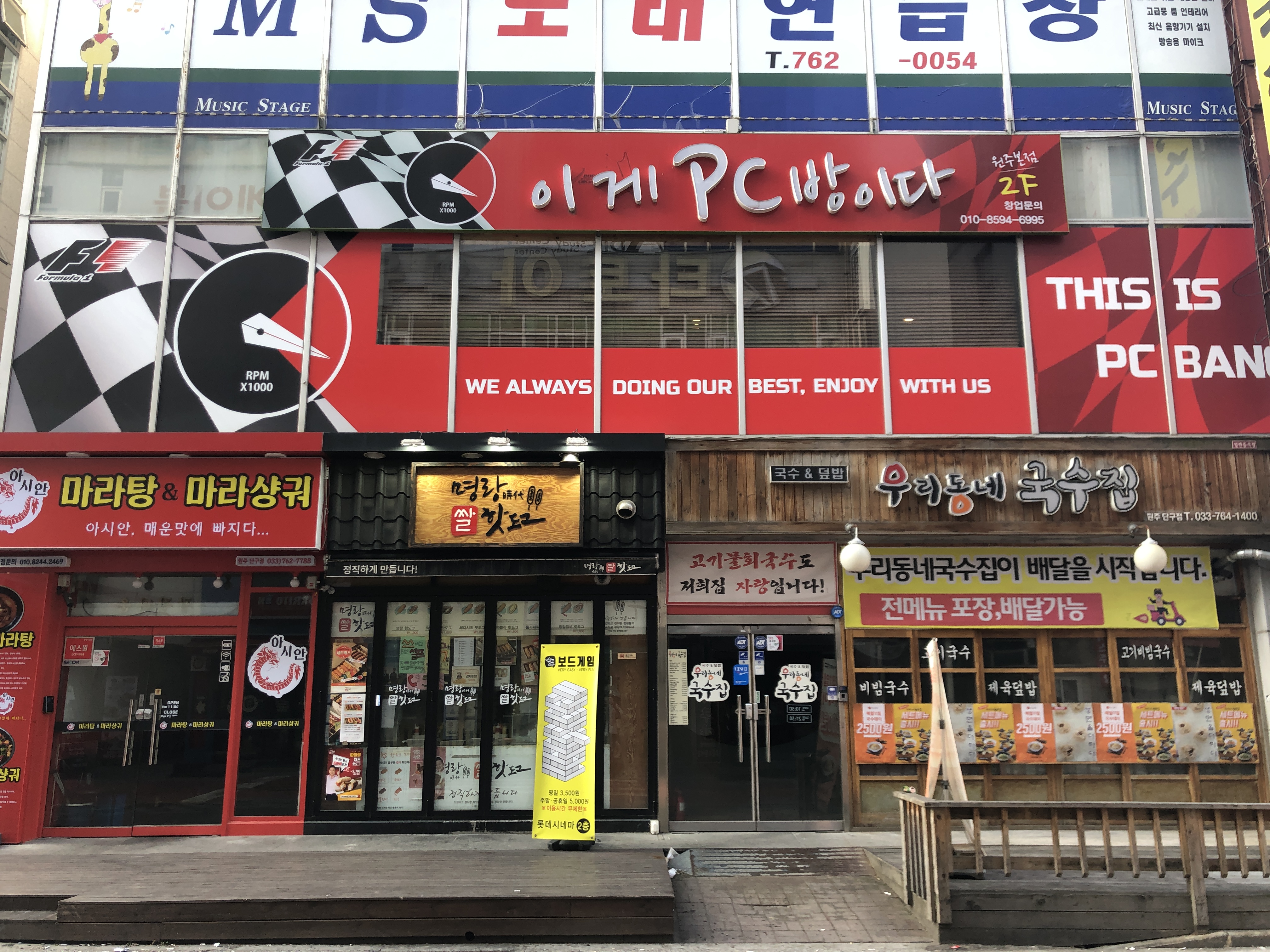 Ein Gebäude mit einer Tafel, auf der 'Das ist PC Bank' steht, in Seoul, Südkorea, mit Glaswänden und -türen, Schildern mit Text und einem Holzzaun davor sowie einer Straße unten.