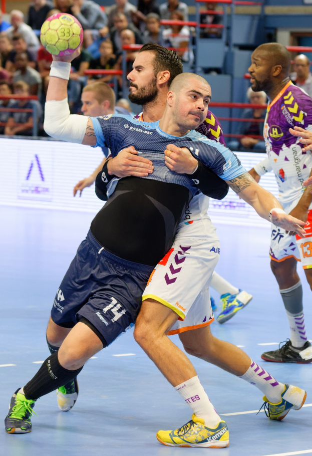 Männer spielen drinnen Handball, einer hält den Ball, mit Zuschauern auf Stühlen, Geländern und einer Wand in einer Halle.