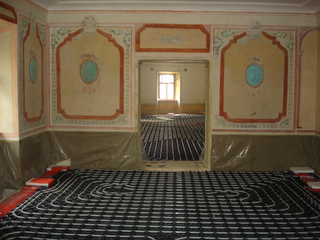 Ein Raum mit einer Fußbodenheizung in der Mitte, umgeben von Wänden und einem Fenster im Hintergrund, mit verschiedenen Gegenständen auf dem Boden, die likely mit dem Heizsystem zusammenhängen.