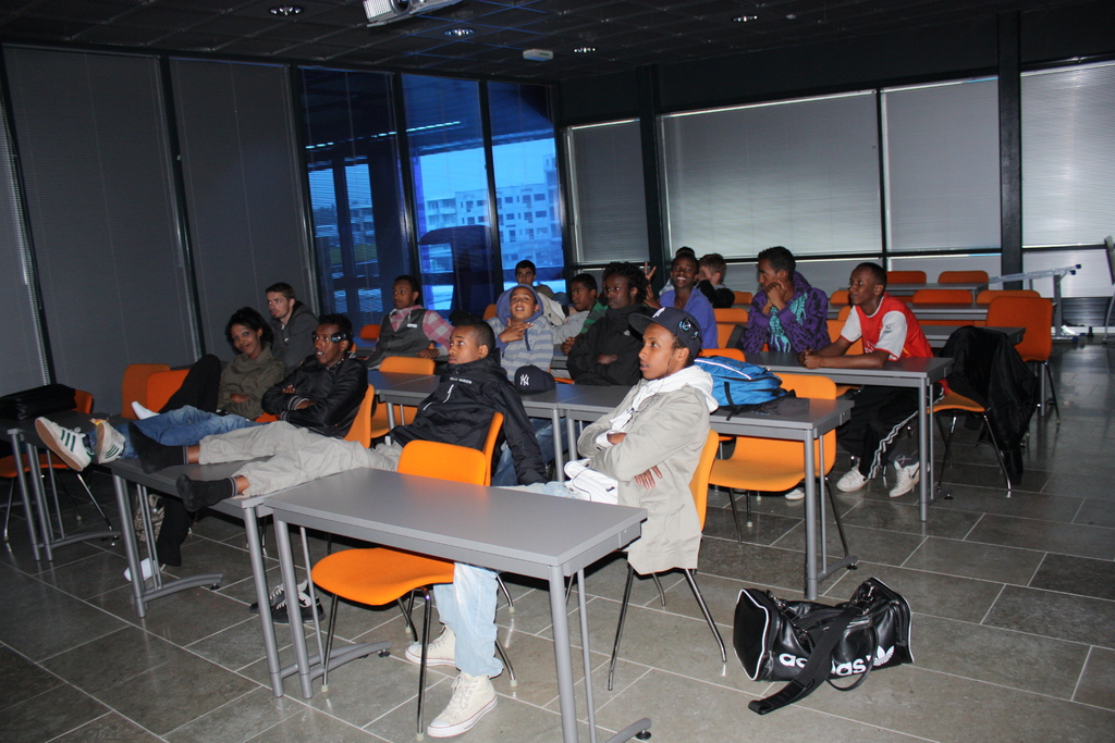 Eine Gruppe von Menschen, die auf Stühlen in einem Klassenzimmer sitzen und auf einen Schreibtisch schauen, mit Fenstern und Vorhängen an der Rückwand.