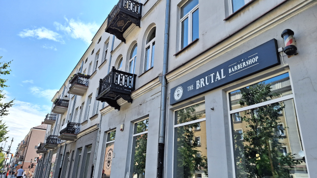 Brutalistischer Barbershop-Laden in Berlin mit Betongebäuden, Fenstern, Geländern, Balkonen und Schildern, Bäumen, Laternenpfählen, Passanten und einer bewölkten Himmel.