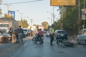 Eine Gruppe von Menschen um ein verunglücktes Motorrad auf der Straße herumsteht, umgeben von mehreren Fahrzeugen, einschließlich eines Lastwagens, und einer Hintergrundlandschaft aus Bäumen, Pfählen, Lampen, Schildern und Himmel.
