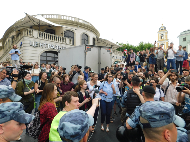 Eine große Gruppe von Menschen steht vor einem Gebäude, einige halten Kameras und Handys, mit einem geparkten Fahrzeug, Bäumen und einem klaren blauen Himmel im Hintergrund, wahrscheinlich protestieren sie gegen das russische Verbot von Drohnen.