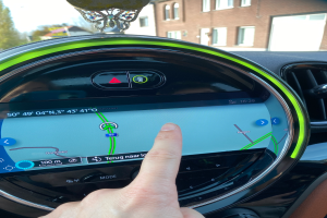 Eine Person zeigt auf ein GPS-Gerät auf dem Armaturenbrett eines Autos, umgeben von Gebäuden, Bäumen und einem klaren blauen Himmel.