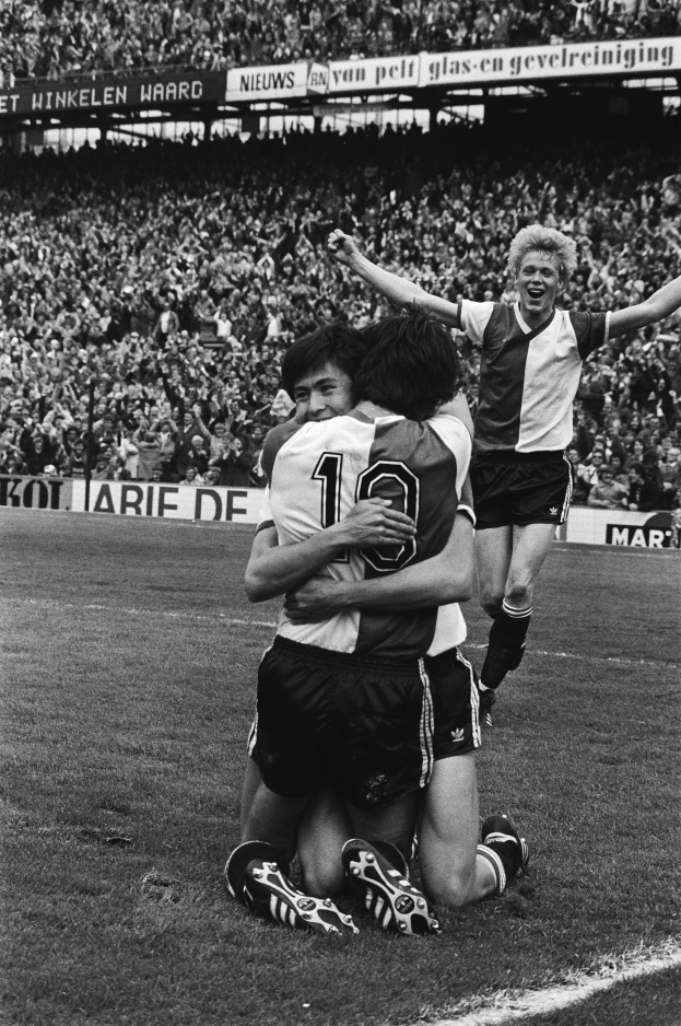 Drei Fussballspieler feiern ein Tor, indem sie sich auf dem Boden umarmen, vor einem jubelnden Publikum und schwenkenden Fahnen im Hintergrund, dargestellt in Schwarz-Weiss.