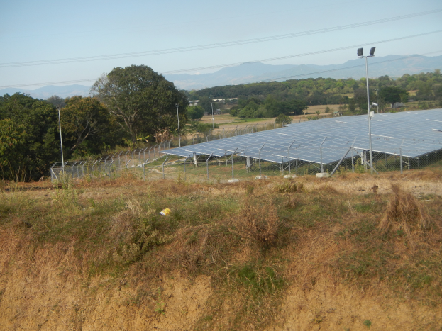 Ein Feld von Solarpanelen auf einem Hügel, umgeben von Gras, Pflanzen, einem Zaun, Pfählen, Lampen, Drähten und Bäumen, mit Hügeln und einem klaren blauen Himmel im Hintergrund.