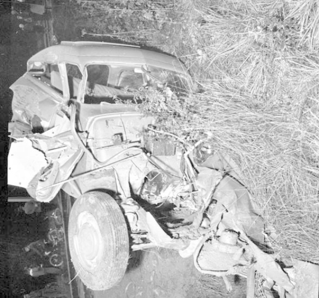 Schwarz-weißes Foto eines schwerbeschädigten Autos mit eingedellter Front und zersplitterten Fenstern am Straßenrand, mit einem Baum und verstreuten Gegenständen im Hintergrund.