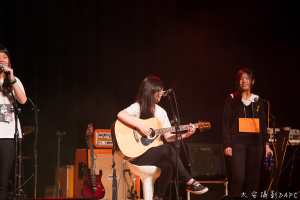Drei Mädchen auf der Bühne, zwei singen in Mikrofone und eine hält ein Musikinstrument, mit einer Gitarre, einem Lautsprecher und einer Schalttafel.