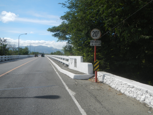 Eine Fahrbahn mit Fahrzeugen, ein Tempolimitschild, Geländer, Laternen, Bäume und ein bewölkter Himmel.