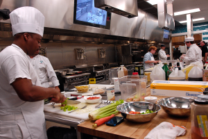 Eine Gruppe von Köchen in einer Küche beim Zubereiten von Speisen, wobei ein Koch im Vordergrund in einer weißen Schürze und Mütze steht und ein Messer hält und von verschiedenen Utensilien, Schüsseln, Gemüse, einem Handy, Flaschen und anderen Gegenständen umgeben ist, während im Hintergrund ein Fernseher und Deckenleuchten zu sehen sind.