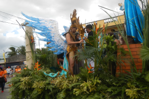 Eine Gruppe von Menschen geht eine Straße entlang neben einem blumengeschmückten Karnevalswagen mit kostümierten Personen, mit Gebäuden, Bäumen, Strommasten und einem bewölkten Himmel im Hintergrund.