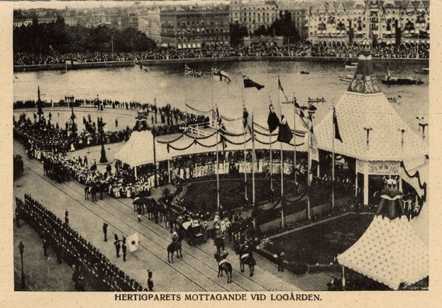 Ein altes Schwarz-Weiß-Foto einer Menge, die sich um einen Rummelplatz mit Zelten, Fahnen, Stangen und Pferden im Vordergrund und Gebäuden, Bäumen und Wasser im Hintergrund versammelt hat, mit Text am unteren Bildrand.