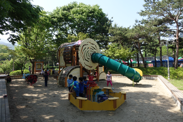 Eine Gruppe von Kindern spielt auf einem Spielplatz mit einer Rutsche in der Mitte, umgeben von Sand und Bäumen, mit einer hölzernen Plattform links, Zelten, Laternenmasten und einem klaren blauen Himmel im Hintergrund.