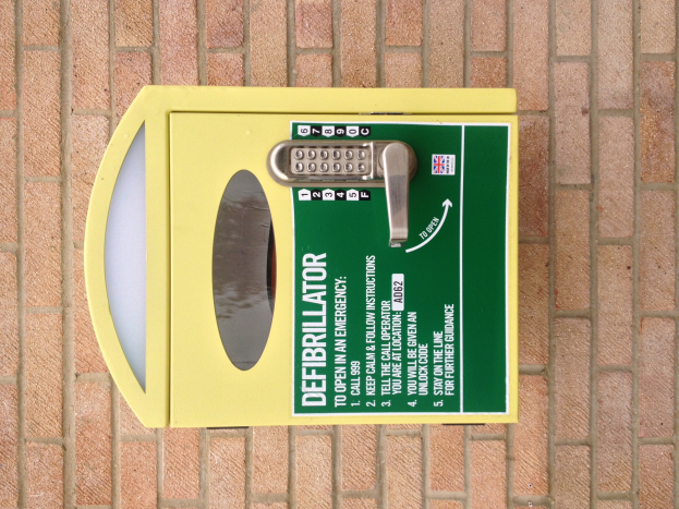 Ein gelber Defibrillator-Kasten mit einer Tastatur oben drauf, der auf einem Ziegelboden steht und bedruckte Texte und Zahlen enthält.