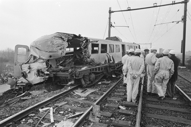 Ein Schwarz-Weiß-Bild einer Zugkollision mit Menschen in Helmen drumherum, Strommasten, Bäumen, Gebäuden und einem klaren Himmel im Hintergrund.